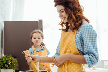 curious daughter looking at mother in polka dot yellow apron putting raw spaghetti in pot