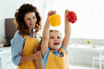 happy mother looking at excited daughter holding red and yellow bell peppers