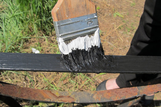 A black paint brush paints a rusty metal railing on a sunny day.