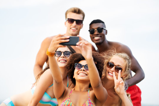 friendship, summer holidays and people concept - group of happy friends taking picture by smartphone on beach