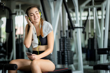 Fototapeta na wymiar Young attractive woman caucasian sitting and listening to music by earphones connect to smart phone. Relaxation after hard workout in gym. Fitness concept, Healthy, Sport, Lifestyle