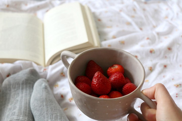 Feet in knit grey socks, bowl of strawberries and book on a bed. Unrecognizable person enjoying leisure time. Selective focus.