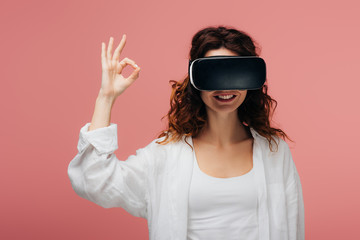 happy curly redhead woman wearing virtual reality headset and showing ok sign on pink