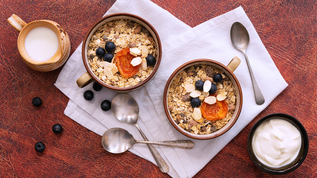 Breakfast setting, two servings of natural bircher muesli with dried apricots; blueberries and slithered almonds, a jug of milk and a bowl of yoghurt. - Powered by Adobe