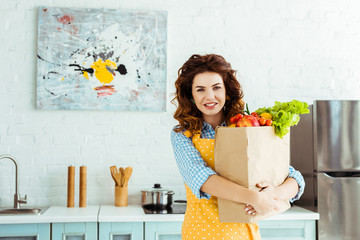 smiling woman in polka dot apron holding paper bag with fresh fruits and vegetables