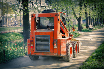 Orange bulldozer with a bucket moves along the path in the city park