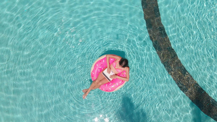 Young female freelancer sitting with a tablet in an inflatable circle in the pool. Aerial view