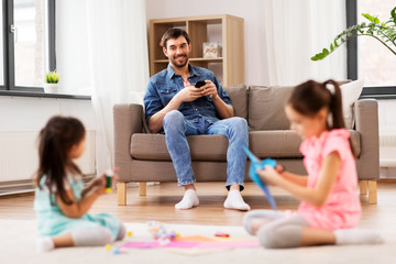 family, leisure and people concept - happy father with smartphone sitting on sofa and looking at his daughters doing crafts at home
