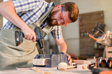 Bearded joiner in safety glasses drills an electric drill hole in a wooden Board in a home workshop repairing wooden products