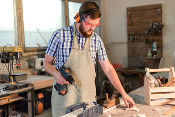 Bearded joiner in safety glasses drills electric drill hole in a wooden Board in the home workshop...