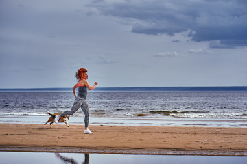 A red-haired middle-aged woman in sportswear runs along the sandy shore of a large river with her...
