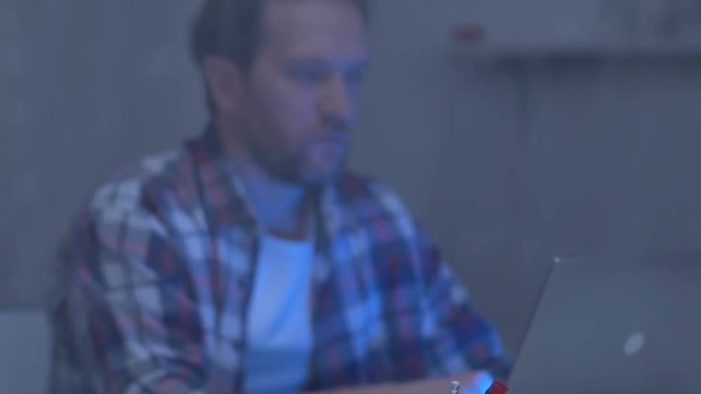 Pensive male freelance developer working on laptop at home behind rainy window