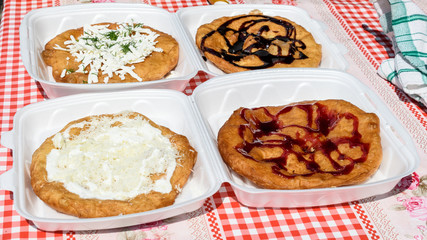 Langos, typical Hungarian food specialty, with sour cream, cheese, dill, chocolate syrup, jam on plastic plates for take away on a table, four deep fried donuts in direct sunlight at a food market