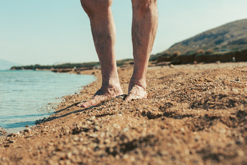 Close up of male hairy legs on yellow sandy beach with sea and hill on background. Male footsteps in the sand. Man standing by the beach. Vacation and Travel concept. Enjoying the summer and sea.