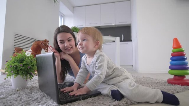modern childhood, happy toddler boy with young mother push buttons laptop computer and clap hands lying on floor indoors