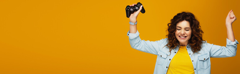 panoramic shot of cheerful curly redhead girl gesturing while holding joystick on orange