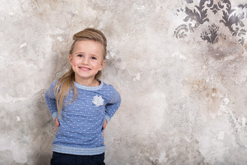 Portrait of a little attractive smiling girl in a blue sweater and pants with hair folded into her hair against a grunge wall background.