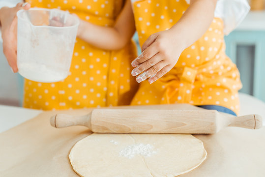 Cropped View Of Mother And Daughter In Yellow Polka Dot Aprons Holding Measuring Jug With Flour Near Dough And Wooden Rolling Pin On Baking Parchment Paper