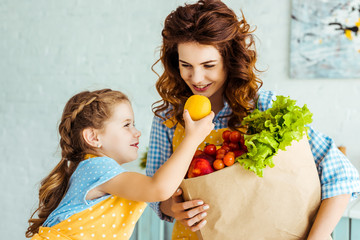smiling daughter giving lemon to mother with paper bag full of ripe fruits and vegetables