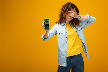 redhead woman covering eyes and holding smartphone with booking app on screen on orange