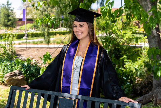 Beautiful young female student standing behind campus bench with arms spread across rail.