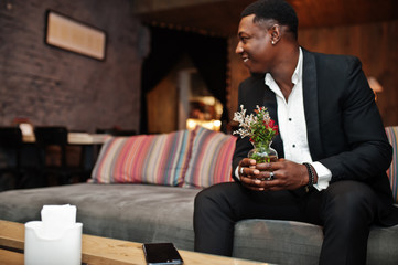 Strong powerful african american man in black suit sitting on couch and hold vase with flowers indoor relax room.