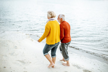 Lovely senior couple dressed in colorful sweaters walking on the sandy beach, enjoying free time during retirement near the sea. Rear view