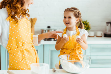excited emotional daughter holding eggs while mother in yellow polka dot apron pointing with finger in kitchen