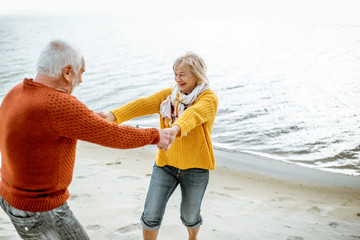Lovely senior couple dressed in colorful sweaters dancing on the sandy beach, enjoying free time during retirement near the sea