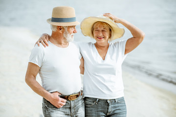 Happy senior couple dressed in white t-shirts and hats walking together on the sandy beach during...