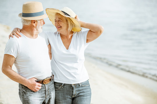 Happy senior couple dressed in white t-shirts and hats walking together on the sandy beach during their retirement
