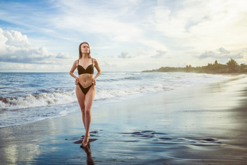 Slim girl in a black swimsuit is standing on a beach against the backdrop of a beautiful sunset.