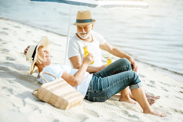 Happy senior couple relaxing, lying with drinks on the sandy beach, enjoying their retirement near the sea