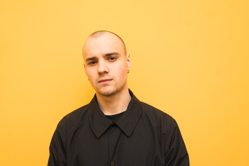 Fototapeta premium Stylish young man with a bald head wearing black jacket poses on a camera on a yellow background and looks seriously in the camera. Trendy authentic guy is isolated on yellow.