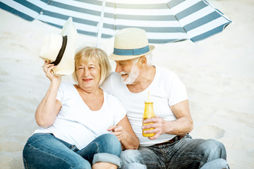 Happy senior couple relaxing, lying together with drinks under umbrella on the sandy beach, enjoying their retirement near the sea