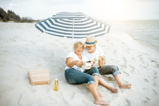 Happy senior couple relaxing, lying together under umbrella on the sandy beach, enjoying their retirement near the sea