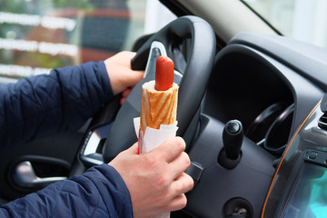 Hot dog in driver hand in car on sunny blurred background. Takeaway, social media. Snack in man...