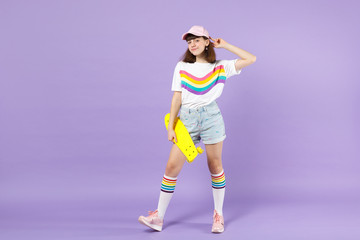Fototapeta na wymiar Portrait of pretty teen girl in vivid clothes standing, holding yellow skateboard isolated on violet pastel wall background in studio. People sincere emotions, lifestyle concept. Mock up copy space.
