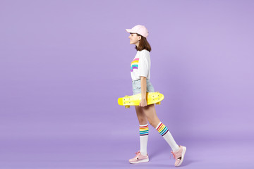 Side view of beautiful cute teen girl in vivid clothes holding yellow skateboard isolated on violet pastel wall background in studio. People sincere emotions, lifestyle concept. Mock up copy space.