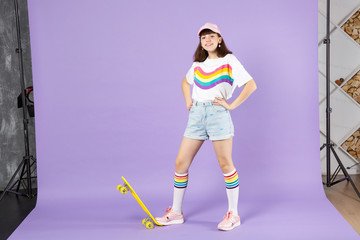 Smiling teen girl in vivid clothes standing with arms akimbo with yellow skateboard isolated on violet pastel wall background in studio. People sincere emotions, lifestyle concept. Mock up copy space.