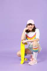 Smiling teen girl in vivid clothes sitting holding yellow skateboard put hand prop up on chin isolated on violet pastel wall background. People sincere emotions, lifestyle concept. Mock up copy space.