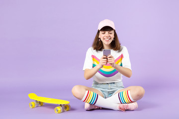 Happy teen girl in vivid clothes sitting near skateboard, using mobile phone, typing sms message isolated on violet pastel background. People sincere emotions, lifestyle concept. Mock up copy space.