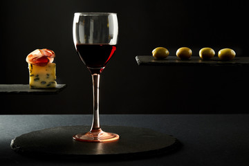 Composition of red wine in glass and pieces of prosciutto and cheese and olives on black background