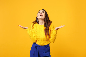 Funny cheerful young woman in sweater, blue trousers looking up spreading pointing hands aside isolated on yellow orange wall background. People sincere emotions lifestyle concept. Mock up copy space.