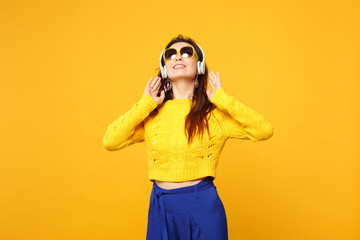 Beautiful young woman in sweater, blue trousers, sunglasses listen music with headphones looking up isolated on yellow orange background. People sincere emotions lifestyle concept. Mock up copy space.