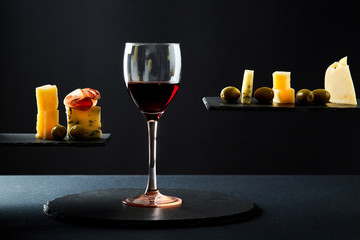 Composition of red wine in glass and pieces of prosciutto and cheese and olives on black background