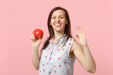 Smiling young woman in summer clothes showing OK gesture holding fresh ripe red apple fruit isolated on pink pastel wall background. People vivid lifestyle, relax vacation concept. Mock up copy space.