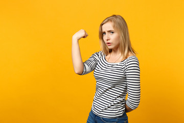 Portrait of displeased young woman in striped clothes looking camera, showing biceps, muscles isolated on yellow orange wall background. People sincere emotions, lifestyle concept. Mock up copy space.