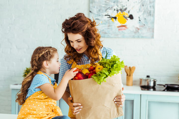happy mother showing curious daughter paper bag with ripe fruits and vegetables