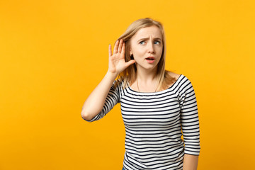 Portrait of curious young woman in striped clothes trying to hear you with hand near ear isolated...